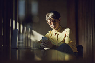 Businesswoman sitting in her sustainable office, using smartphone - MCF00648