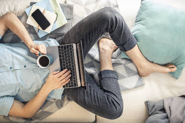 Blick von oben auf einen Mann, der mit einer Tasse Kaffee und einem Laptop auf dem Sofa liegt - MSUF00270