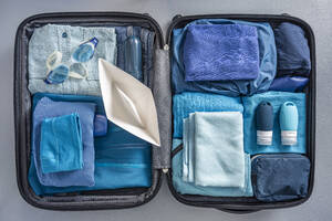 Draufsicht auf einen gepackten Koffer mit Kleidung, Schwimmbrille und Papierboot - MSUF00288