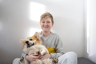 Portrait of boy sitting on bed at home cuddling his dog - VPIF02319