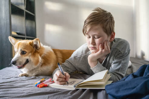 Porträt eines zeichnenden Jungen, der mit seinem Hund auf dem Bett liegt - VPIF02321