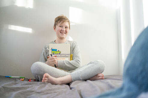 Porträt eines glücklichen Jungen, der auf einem Bett sitzt und eine Zeichnung zeigt - VPIF02327