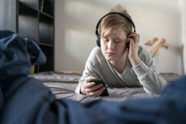 Porträt eines Jungen mit Kopfhörern, der auf dem Bett liegt und auf sein Handy schaut - VPIF02331