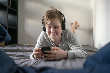 Portrait of smiling boy with headphones lying on bed looking at cell phone - VPIF02332