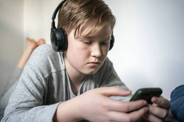 Portrait of serious boy lying on bed with headphones looking at cell phone - VPIF02333