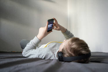 Boy with headphones lying on bed using cell phone - VPIF02334