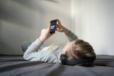 Junge mit Kopfhörern, der auf dem Bett liegt und ein Mobiltelefon benutzt - VPIF02334