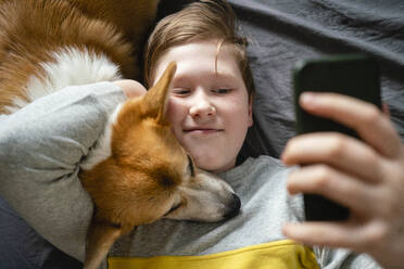 Portrait of boy lying on bed with his dog taking selfie with smartphone - VPIF02336