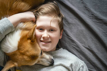 Portrait of smiling boy lying on bed cuddling his dog - VPIF02337