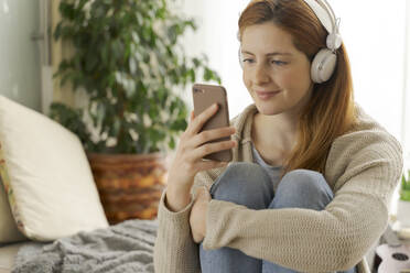 Junge Frau trägt Kopfhörer und benutzt ihr Smartphone zu Hause - AFVF05991