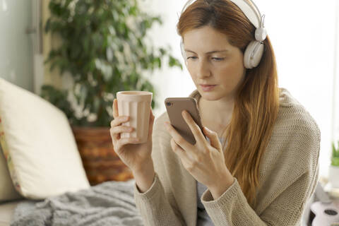 Junge Frau trägt Kopfhörer und benutzt ihr Smartphone zu Hause, lizenzfreies Stockfoto