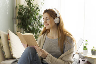 Junge Frau trägt Kopfhörer und liest zu Hause in einem Notebook - AFVF05995