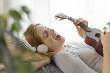 Unbekümmerte junge Frau mit Kopfhörern und Ukulele zu Hause - AFVF06026