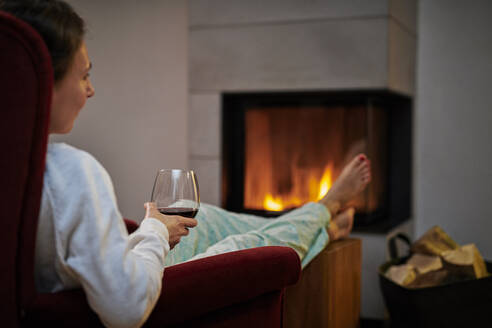 Frau sitzt auf einem Sessel mit einem Glas Rotwein und entspannt sich vor einem Kamin - DIKF00452