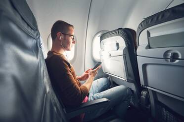 Mann hört Musik, während er sein Mobiltelefon im Flugzeug benutzt - EYF04599