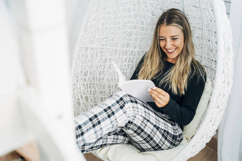 Portrait of laughing woman sitting on swinging chair at home reading a book - MPPF00776