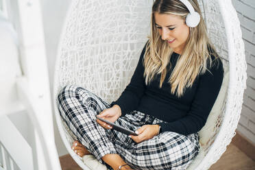 Lächelnde Frau, die zu Hause auf einem Schaukelstuhl sitzt und Kopfhörer und ein Mini-Tablet benutzt - MPPF00782