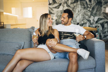 Happy couple sitting on the couch with glasses of white wine - MPPF00799