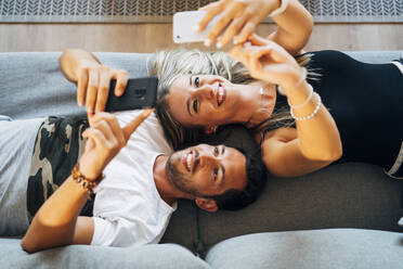 Porträt eines lachenden Paares, das auf der Couch liegt und Selfies mit Smartphones macht - MPPF00803