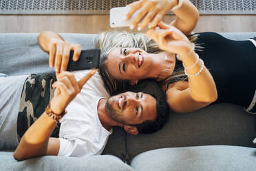 Portrait of laughing couple lying on the the couch taking selfies with smartphones - MPPF00803