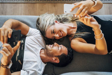 Happy couple lying on the couch taking selfies with smartphones while looking at each other - MPPF00804