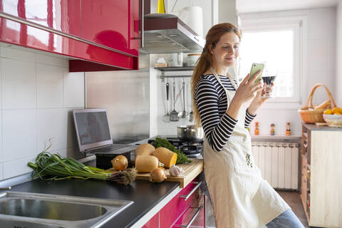 Woman drinking glass of red wine in the kitchen, while using smartphone stock photo