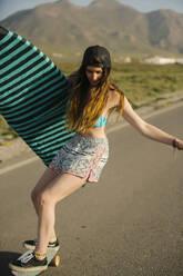 Junge Frau mit Surfbrett, die auf einer asphaltierten Straße Skateboard fährt, Almeria, Spanien - MPPF00807
