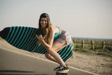 Porträt einer glücklichen jungen Frau mit Surfbrett, die auf einer asphaltierten Straße Skateboard fährt, Almeria, Spanien - MPPF00809