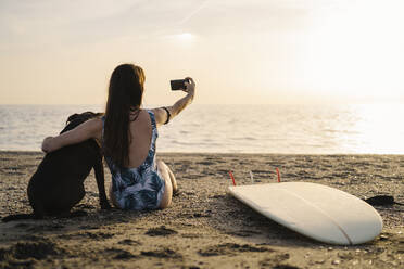 Rückenansicht einer jungen Surferin, die mit ihrem Hund am Strand sitzt und ein Selfie mit ihrem Smartphone macht, Almeria, Spanien - MPPF00828