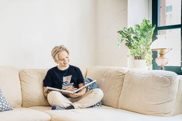 Glücklicher Junge sitzt auf der Couch im Wohnzimmer zu Hause und liest ein Buch - MJF02444