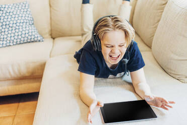 Schreiender Junge auf der Couch zu Hause mit Headset und Tablet - MJF02447