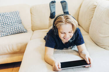 Junge auf der Couch zu Hause mit Headset und Tablet liegend - MJF02448