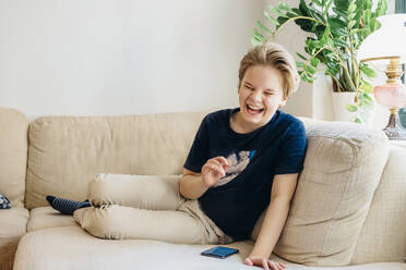 Lachender Junge auf der Couch im Wohnzimmer zu Hause mit Smartphone - MJF02452