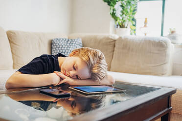 Boy lying on coffee table with smartphone and tablet taking a nap - MJF02458