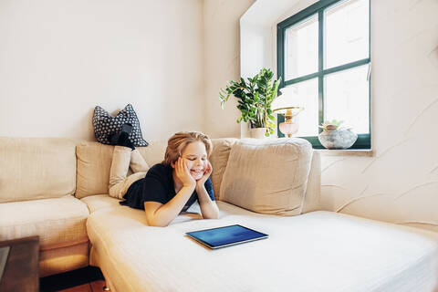 Lächelnder Junge auf der Couch im Wohnzimmer zu Hause mit Tablet, lizenzfreies Stockfoto