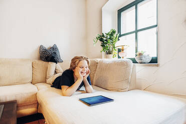 Lächelnder Junge auf der Couch im Wohnzimmer zu Hause mit Tablet - MJF02478