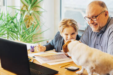 Father helping son doing homework with dog on desk - MJF02503