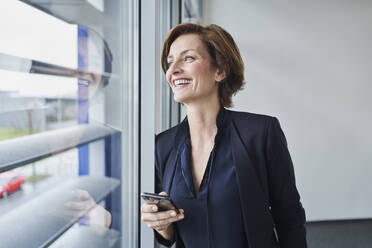 Happy businesswoman with cell phone at the window - RORF02124