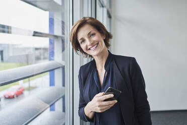 Portrait of confident businesswoman at the window holding cell phone - RORF02125