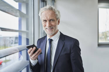 Porträt eines lächelnden Geschäftsmannes, der ein Handy am Fenster hält - RORF02133