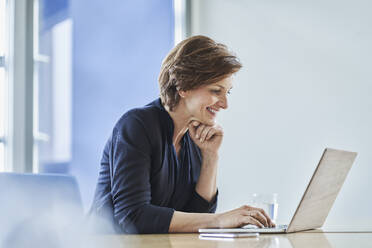 Smiling businesswoman using laptop at desk in office - RORF02141