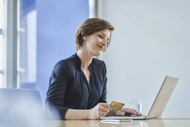 Lächelnde Geschäftsfrau, die eine Kreditkarte hält und einen Laptop am Schreibtisch im Büro benutzt - RORF02145