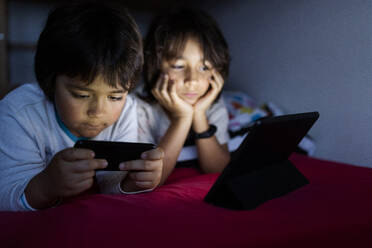 Zwei Brüder liegen zu Hause auf dem Bett und benutzen elektronische Geräte - VABF02781