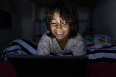 Porträt eines grinsenden Jungen, der zu Hause auf dem Bett liegt und ein digitales Tablet benutzt - VABF02783