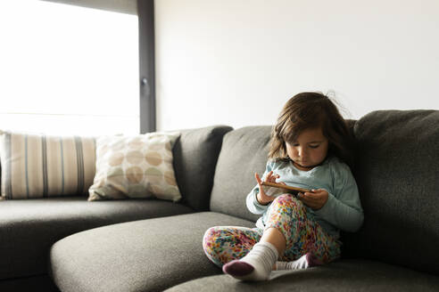 Toddler girl sitting on couch at home using smartphone - VABF02795