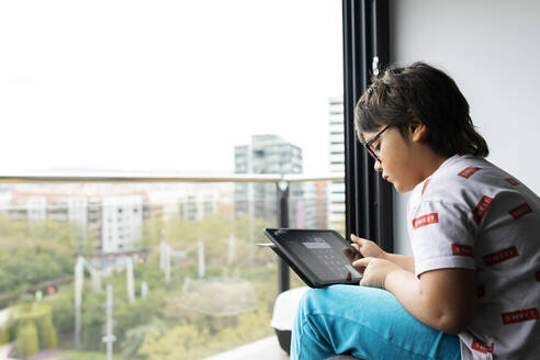 Boy sitting in front of window at home using digital tablet - VABF02799