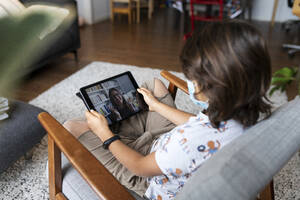 Boy with surgical mask sitting on armchair using digital tablet for video chat with his mother - VABF02811