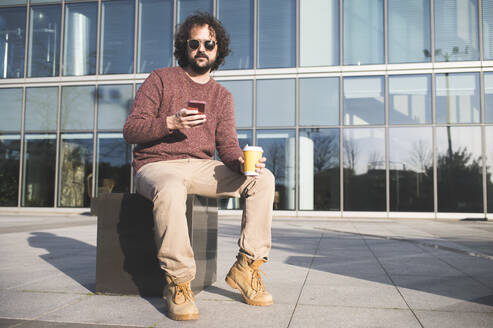 Portrait of bearded man sitting outdoors with coffee to go and smartphone - KIJF02971