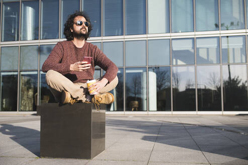 Portrait of bearded man sitting outdoors with coffee to go and smartphone enjoying sunlight - KIJF02972