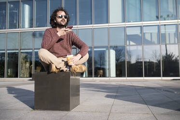 Porträt eines bärtigen Mannes, der mit einem Kaffee zum Mitnehmen im Freien sitzt und telefoniert - KIJF02973
